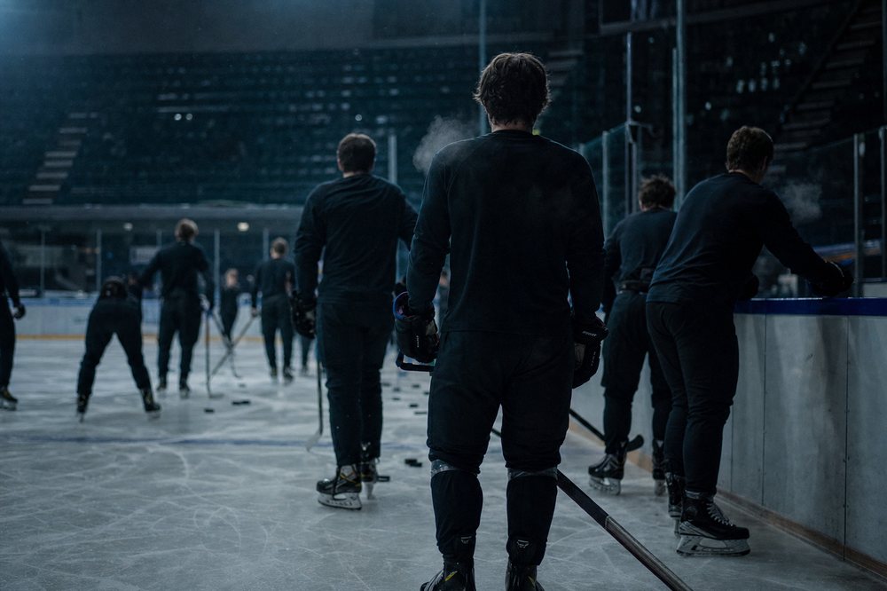 Groep ijshockeyspelers tijdens een teamtraining langs de boarding in een koude, donkere ijshal.