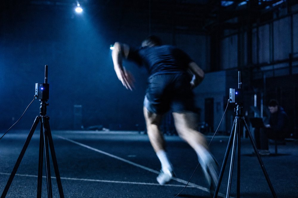 Sprinttest in een donkere sporthal: timing gates op statieven met daartussen een versnellende atleet in beweging.